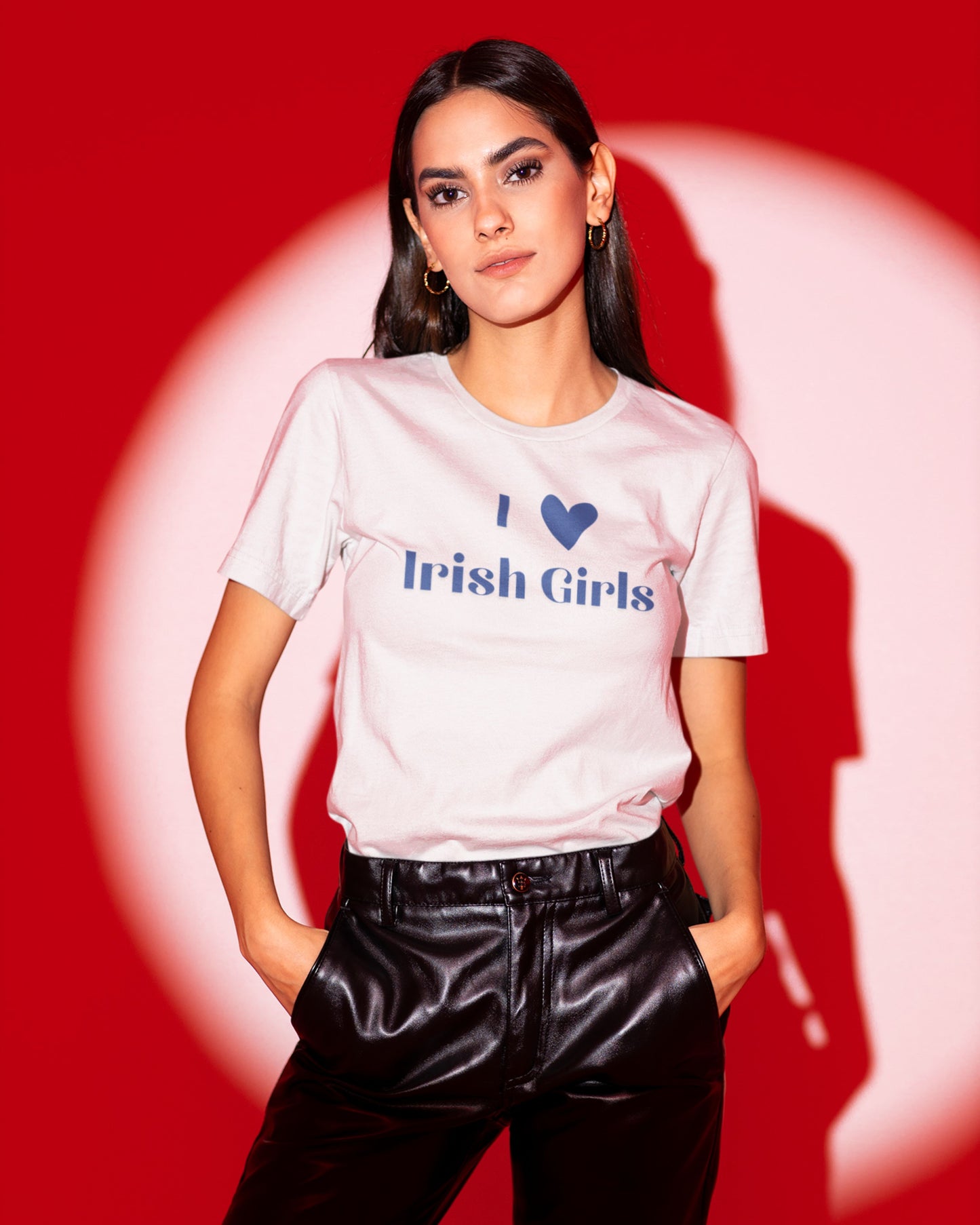 Girl with slick back hair wearing a white shirt that says I heart Irish Girls. This shirt is a St. Patricks Day design. She is wearing leather pants and a red background.